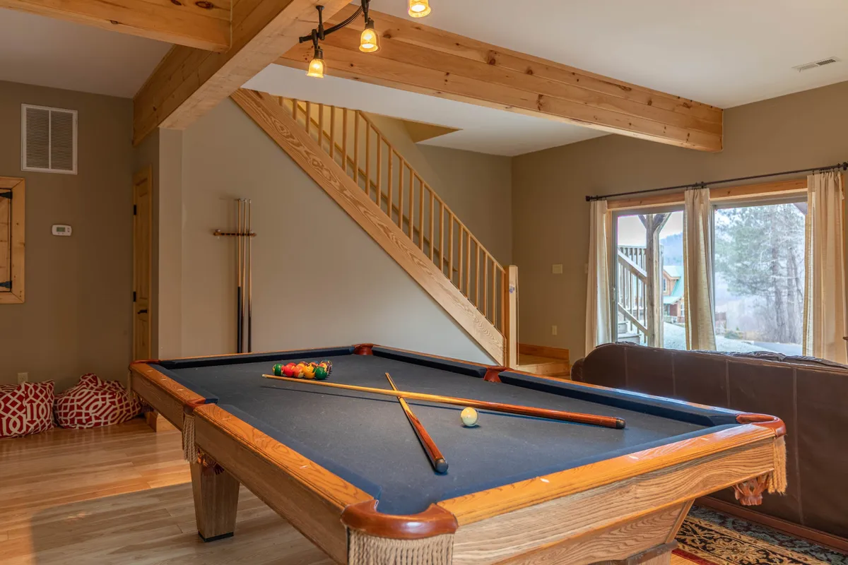 A lively view of the game room showing a pool table, a staircase leading upstairs, and a large window filling the space with light. The setting suggests a dynamic area where games and laughter take center stage.