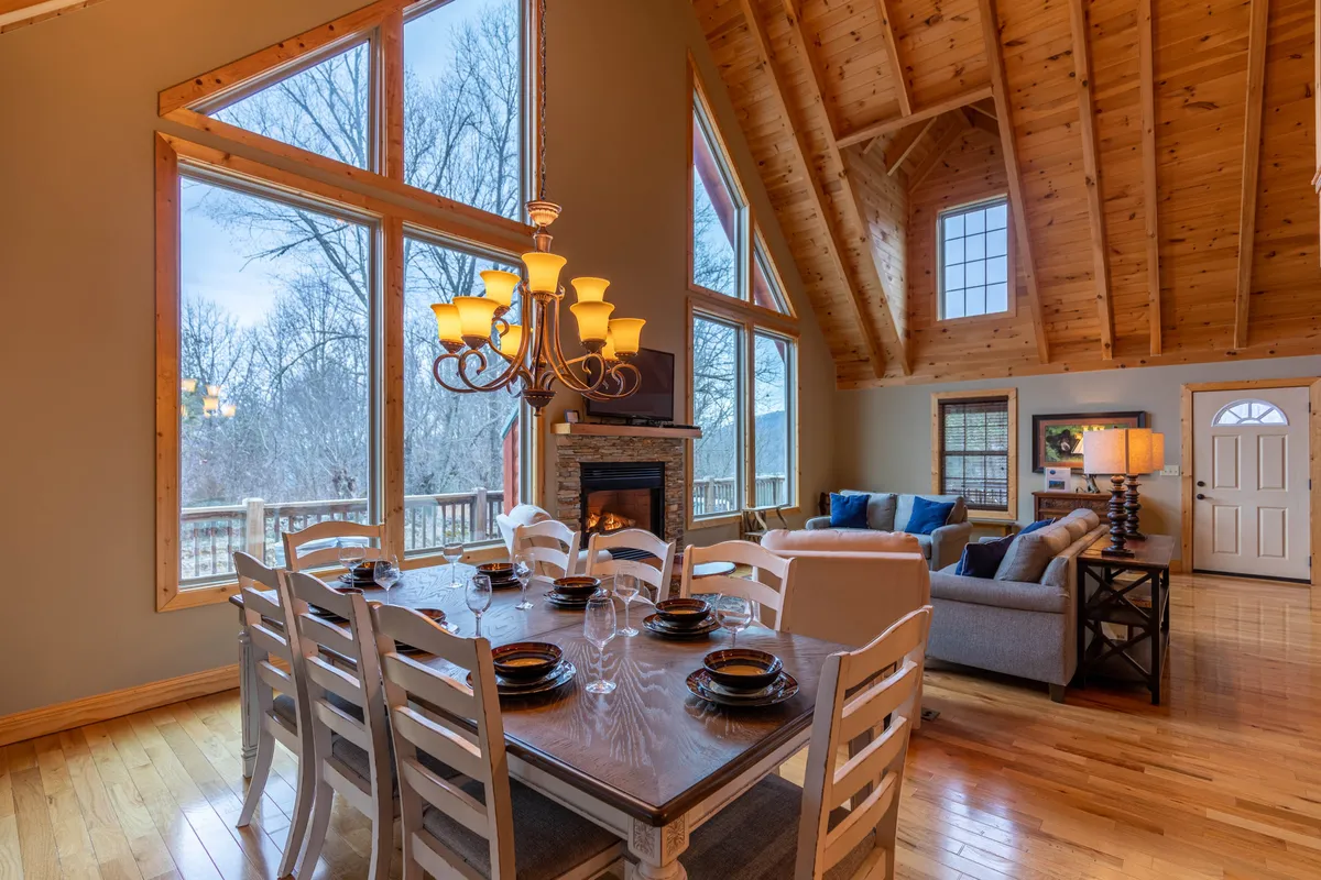 A wide-angle view of the spacious family room showcasing a large dining table set for a gathering, a cozy sofa seating area positioned near a glowing gas log fireplace, and expansive windows that flood the space with soft mountain light. The open layout seamlessly connects the living, dining, and kitchen areas, highlighting the communal atmosphere.