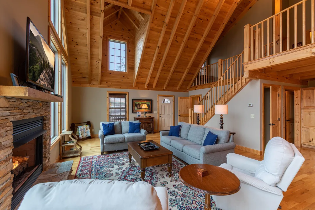 A warm and welcoming living area with a prominent fireplace, large windows revealing natural light, exposed wooden beams, and comfortable seating arranged for relaxation or socializing.