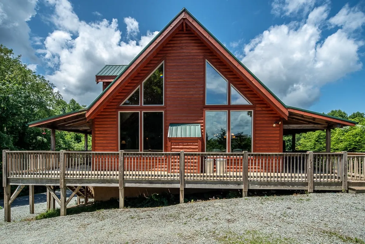 This image captures the exterior of the cabin with its large front windows, inviting wooden deck, and A-frame structure. The deck is set up for gatherings, while the surrounding scenery invites outdoor relaxation and group activities.