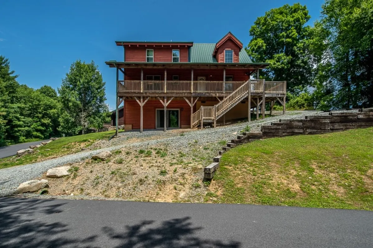 A welcoming exterior shot of the cabin features a spacious wooden deck with a staircase leading to the entrance, a striking red facade with a green roof, and is nestled among tall trees. The setting evokes the sense of being surrounded by nature, with ample space for outdoor gatherings, relaxing, and reconnecting after adventures.