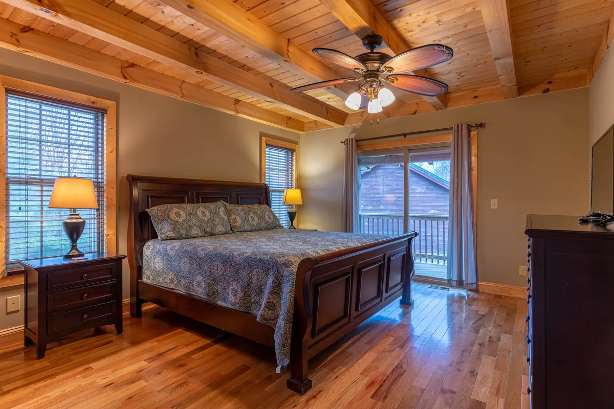 A bright and inviting bedroom with a comfortable bed, nightstand, windows letting in natural light, a lamp, and a dresser—perfect for restful nights or quiet reading.