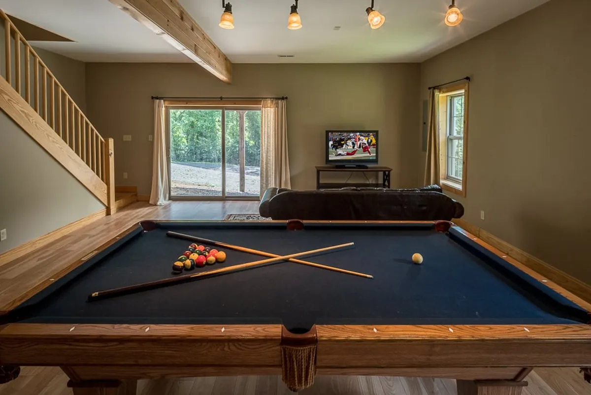 A vibrant game room featuring a pool table in the center, large windows that let in natural light, and a TV mounted on the wall. The space is arranged to encourage both play and relaxation, with comfortable seating positioned for socializing and watching TV.