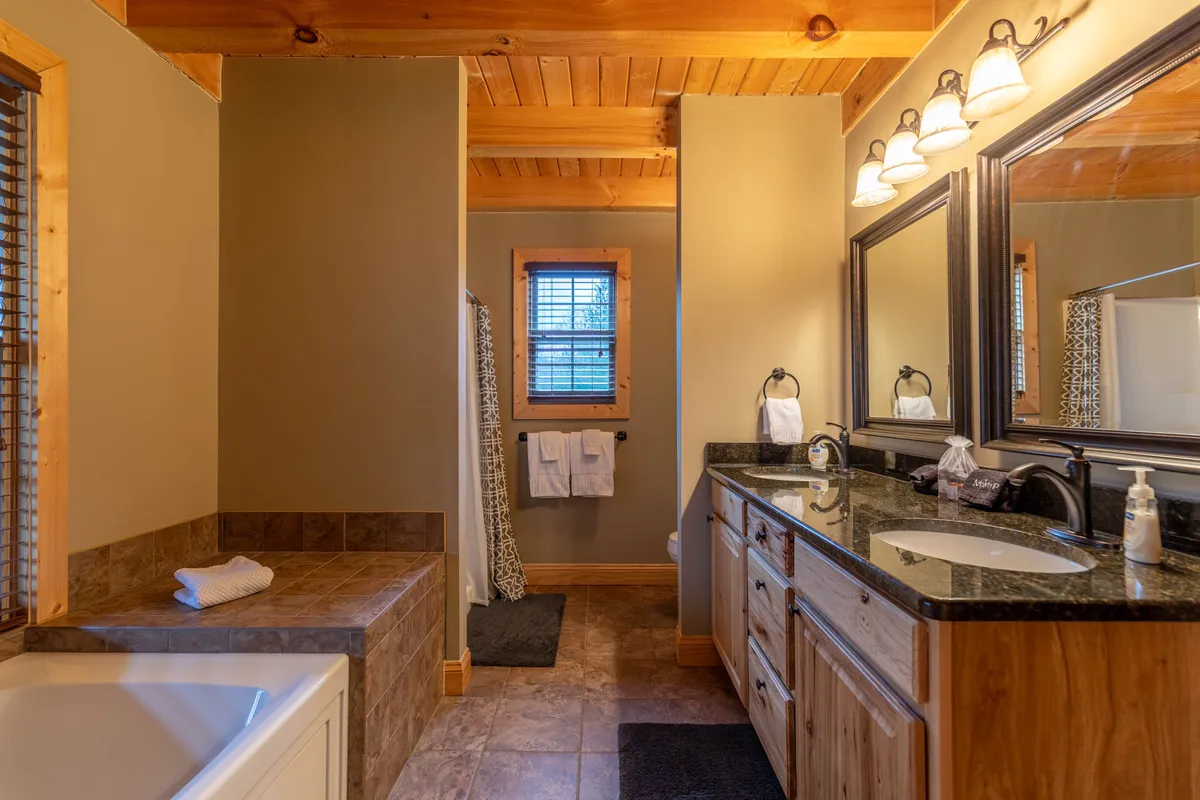A spacious bathroom featuring a large soaking tub, double sink vanity, framed mirrors, wooden ceiling, and a window with blinds, evoking a sense of relaxation and luxury.