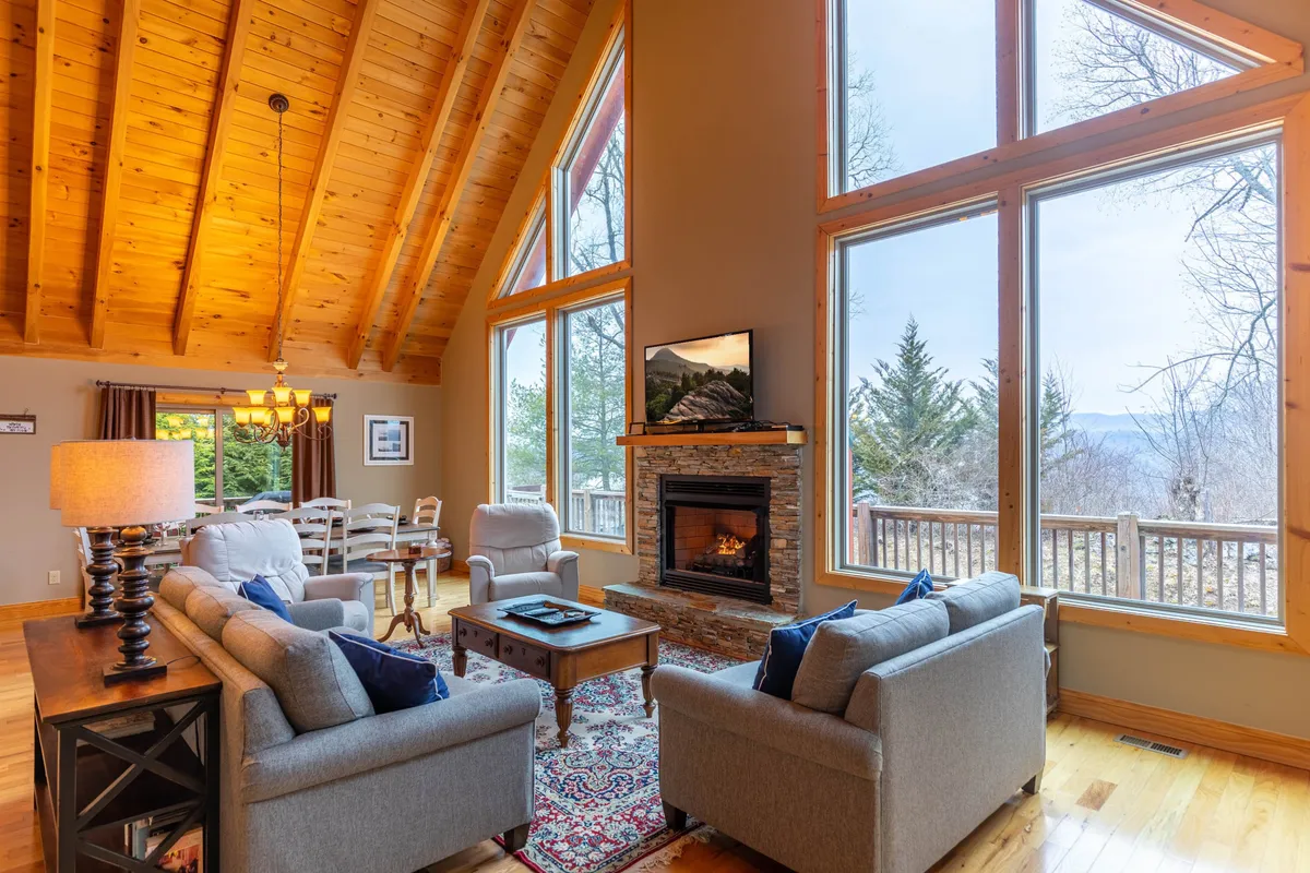 A warm, welcoming interior scene featuring a wood-beamed ceiling, large windows with mountain views, a prominent stone fireplace, and a dining area adjacent to the living space. The image radiates a cozy, family-friendly atmosphere ideal for gatherings.