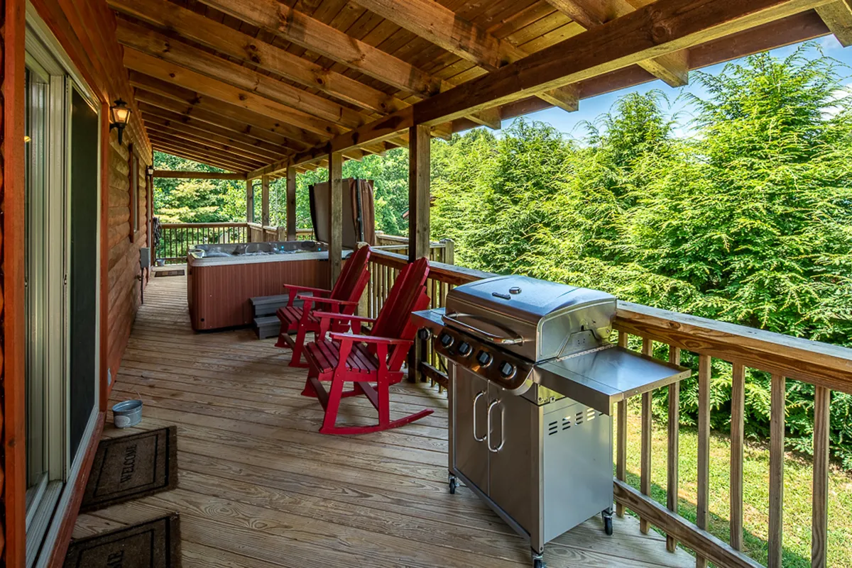 This image showcases a spacious wooden deck featuring red chairs, a hot tub, grill, and lush green trees in the background. The layout demonstrates how the deck seamlessly accommodates both relaxation in the hot tub and outdoor cooking at the grill, while inviting seating encourages social gatherings.