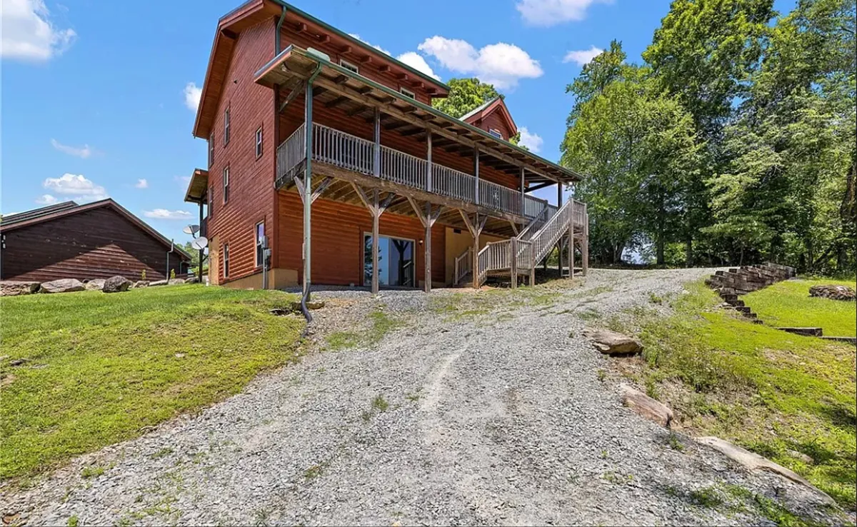 An exterior view of the property focuses on a wooden balcony overlooking a lush green lawn and surrounding forest. The gravel driveway and spacious porch suggest easy access for vehicles and a welcoming space for gathering and relaxing outdoors.