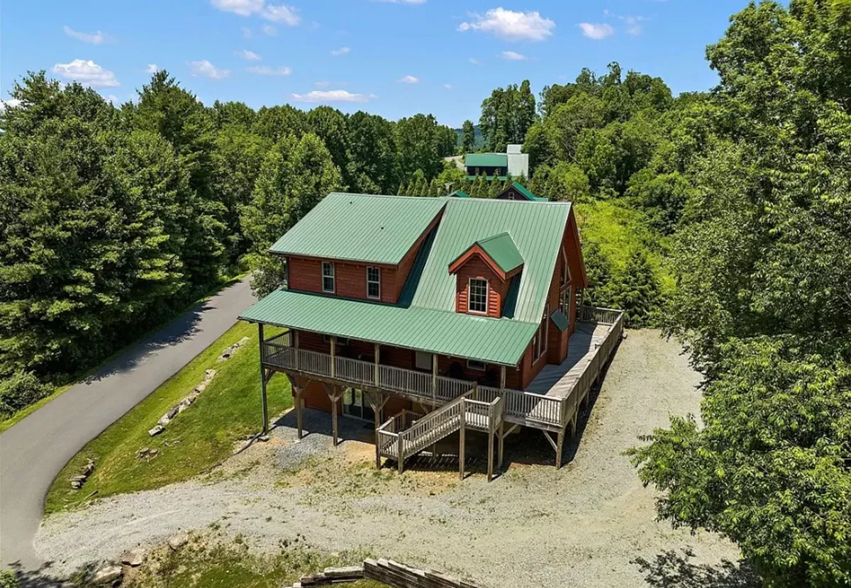 The hero image features a charming log cabin with a classic green metal roof, nestled among lush trees. A spacious wooden deck wraps around the home, perfect for relaxing or entertaining, while the surrounding forest hints at peaceful seclusion. The inviting exterior exudes warmth and promises cozy mountain living.